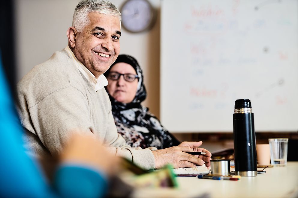 Ein älterer Mann und eine Frau mit Kopftuch sitzen an einem Konferenztisch.