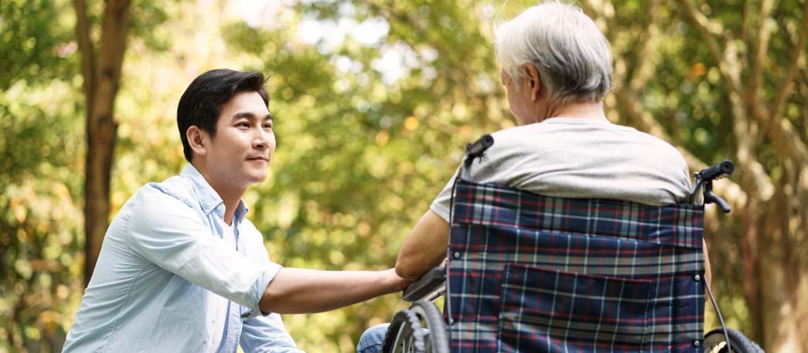 Ein asiatischer Mann kniet neben dem Vater im Rollstuhl im Freien. 